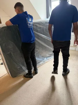 Two SJC Movers carrying a plastic-wrapped divan base out of a bedroom